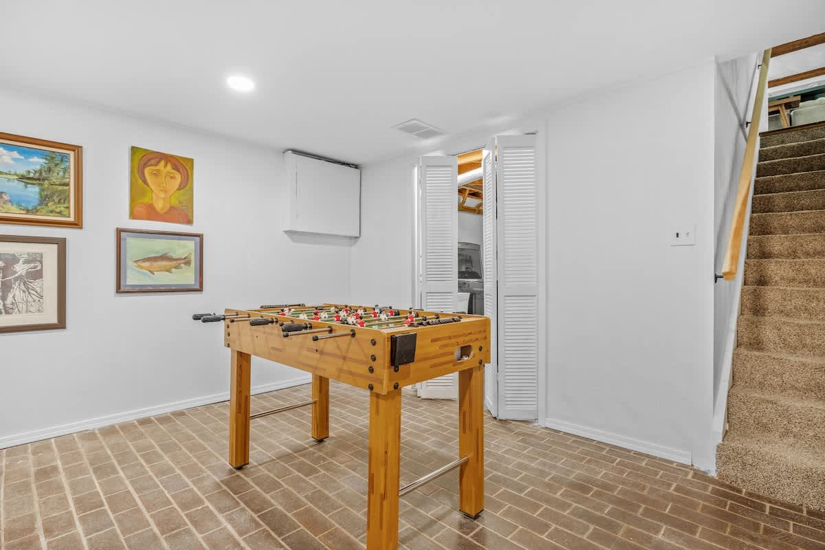 A bright game room features a wooden foosball table on a brick-patterned floor, with white walls adorned with framed artwork, and a staircase leading up on the right.