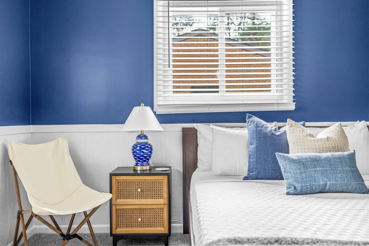 A cozy bedroom with a dark blue accent wall and white wainscoting features a bed with white bedding and blue accent pillows, a black nightstand with a blue and white lamp, and a cream-colored folding chair. A window with white blinds is visible above the nightstand.