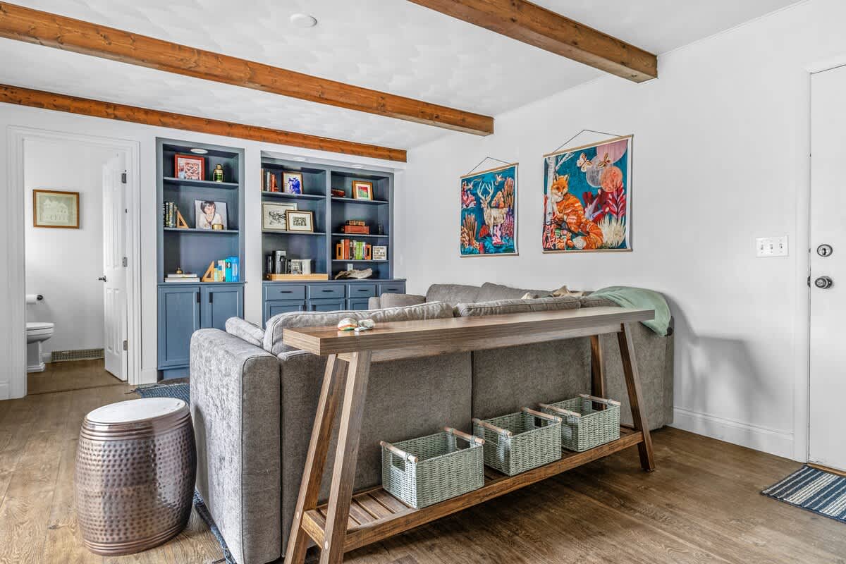 A stylish living room features a gray sectional sofa, a long wooden console table with storage baskets, and built-in blue bookshelves, all under exposed wooden ceiling beams.