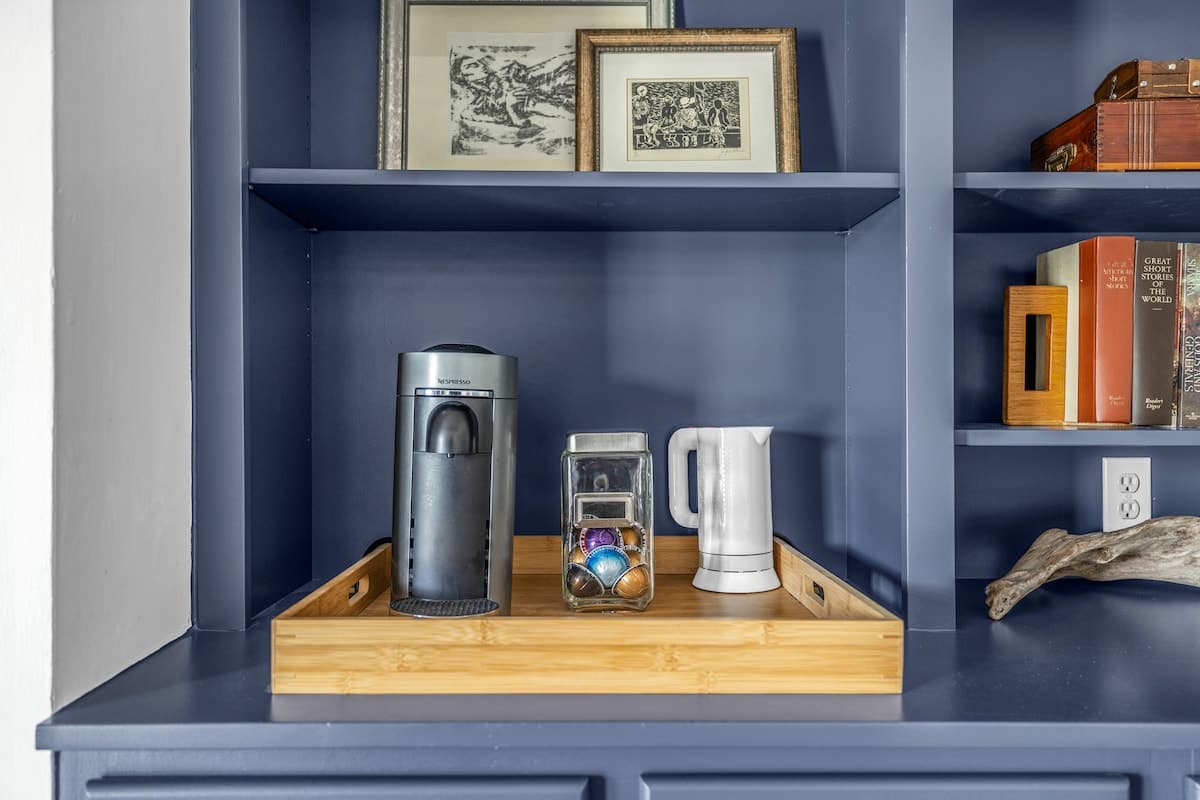 A built-in blue cabinet with open shelving displays a Nespresso machine, a glass jar of coffee pods, and a white electric kettle on a bamboo tray, alongside decorative items and books.