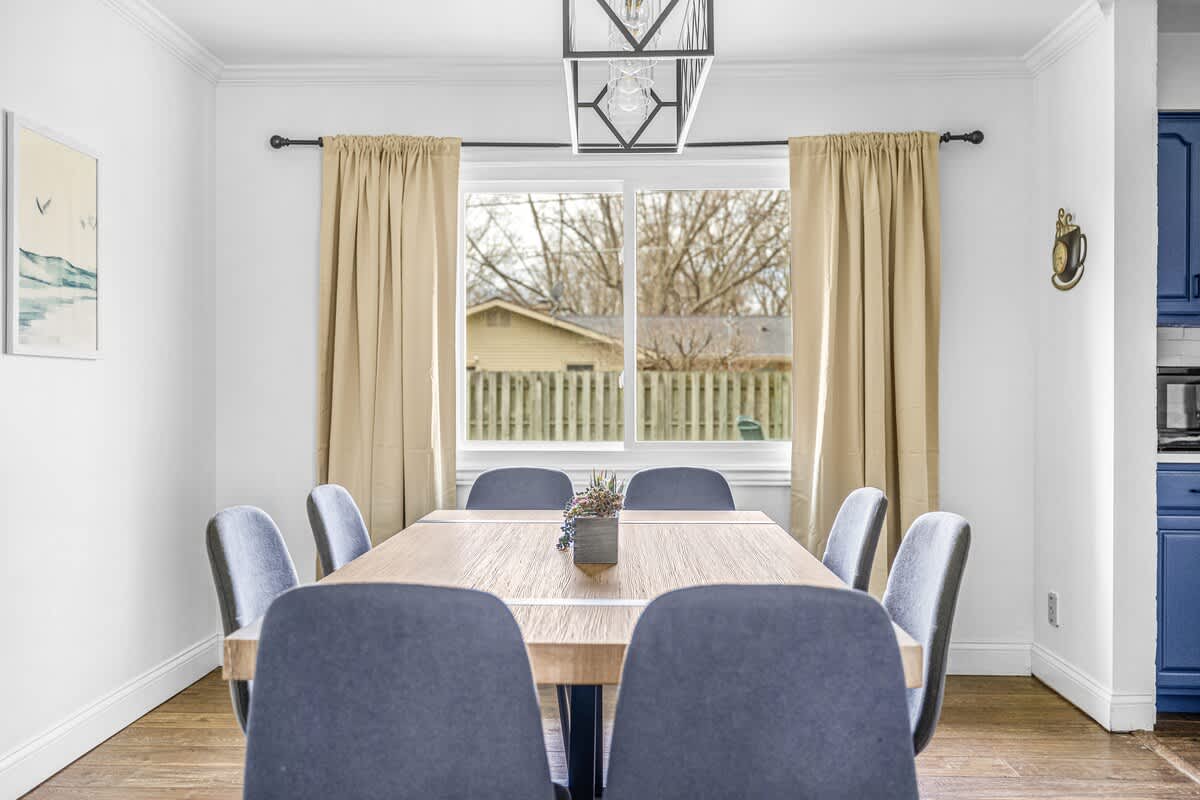 A bright dining room showcases a long wooden table with eight blue upholstered chairs, centered under a modern geometric chandelier, with a large window overlooking a backyard. Light beige curtains frame the window.