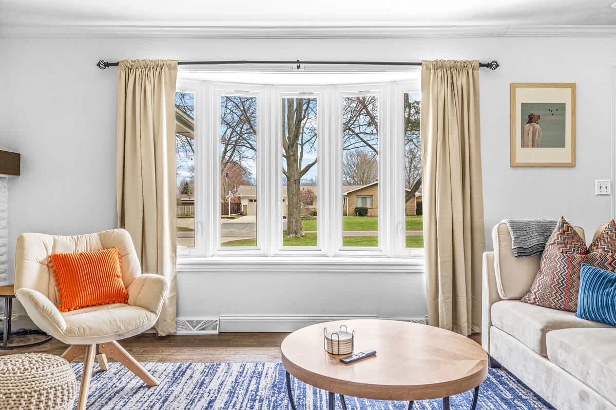 A bright living room features a large bay window with beige curtains, a cream armchair with an orange pillow, and a round wooden coffee table on a blue and white striped rug.
