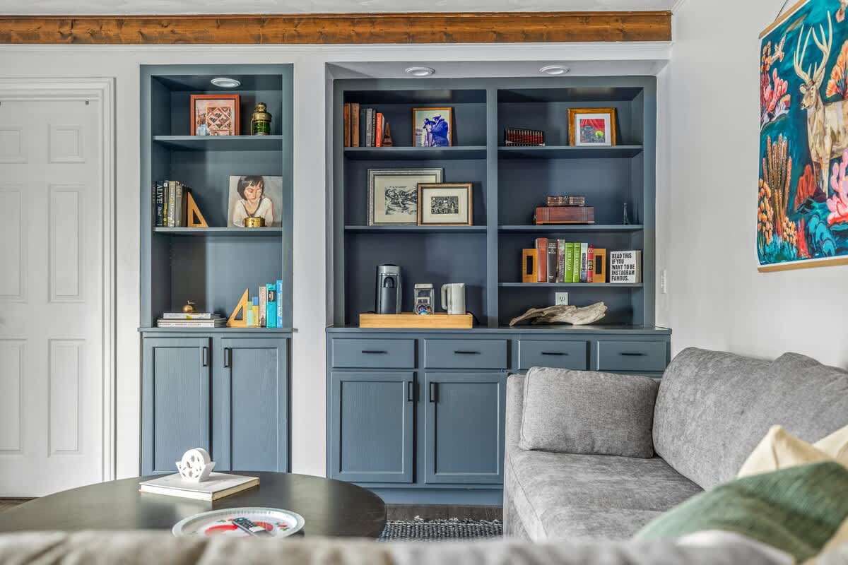 A cozy living space features built-in blue bookshelves and cabinets filled with decor and books, next to a gray sectional sofa, with a dark wood coffee table and colorful wall art, under exposed wooden beams.