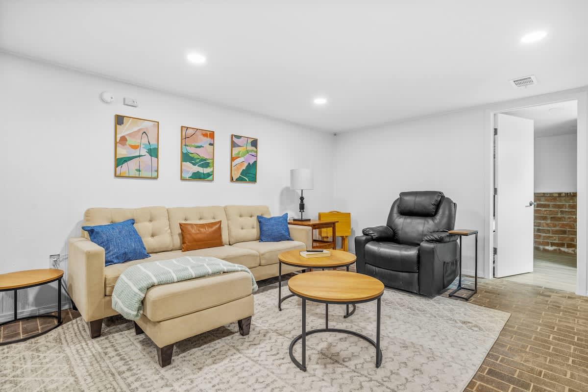 A bright, modern living area with white walls, recessed lighting, a beige sofa with blue and brown throw pillows, and a black recliner. Two round wooden coffee tables sit on a patterned area rug, with three abstract art pieces adorning the wall.
