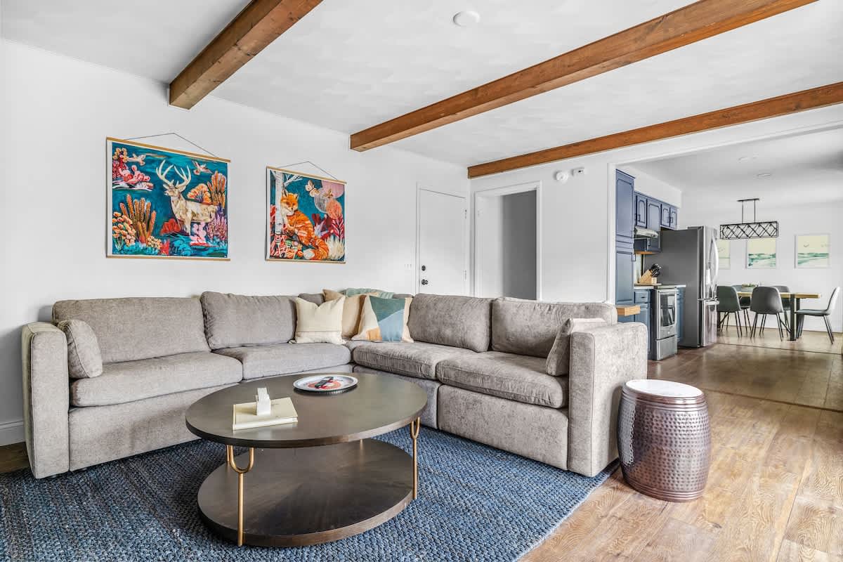 A modern living room features a large gray sectional sofa, a dark oval coffee table, and exposed wooden ceiling beams, with colorful framed art on the white walls and a blue area rug.