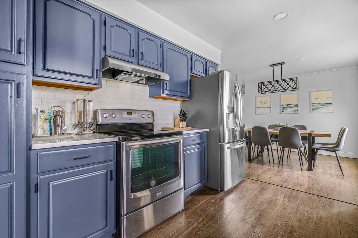 Another view of the galley kitchen, highlighting the stainless steel range and refrigerator, dark blue cabinets, and white countertops, with the dining room visible in the background.