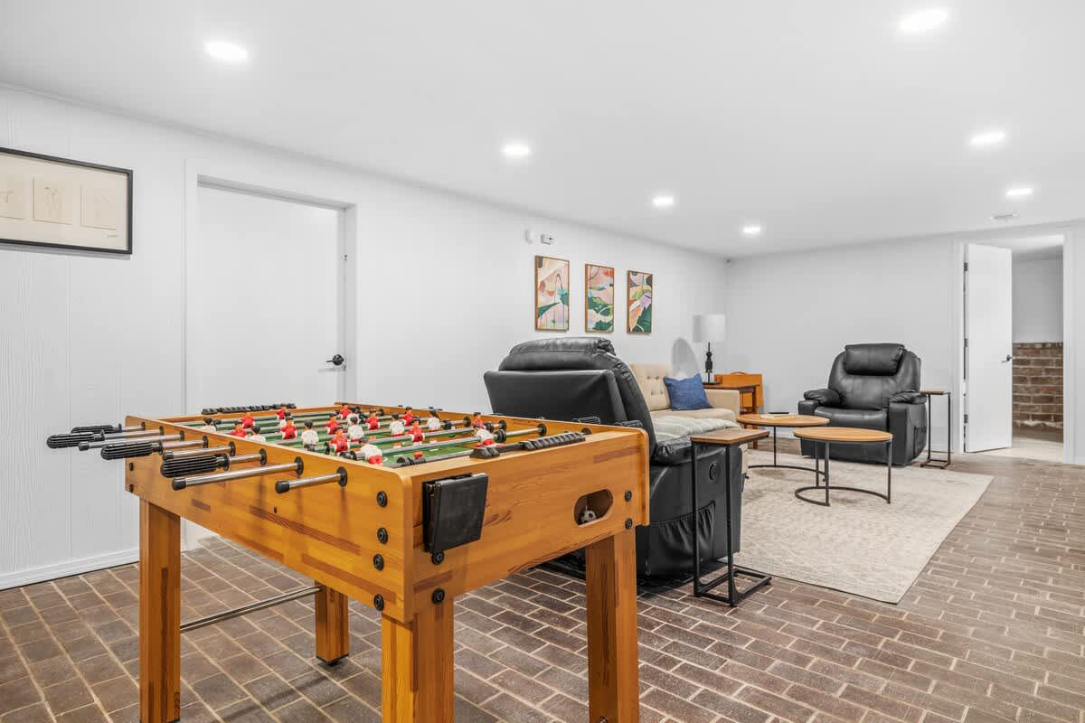 A spacious game room featuring a wooden foosball table in the foreground, with a comfortable living area visible in the background. The room has white walls, recessed lighting, and a brick-patterned floor.