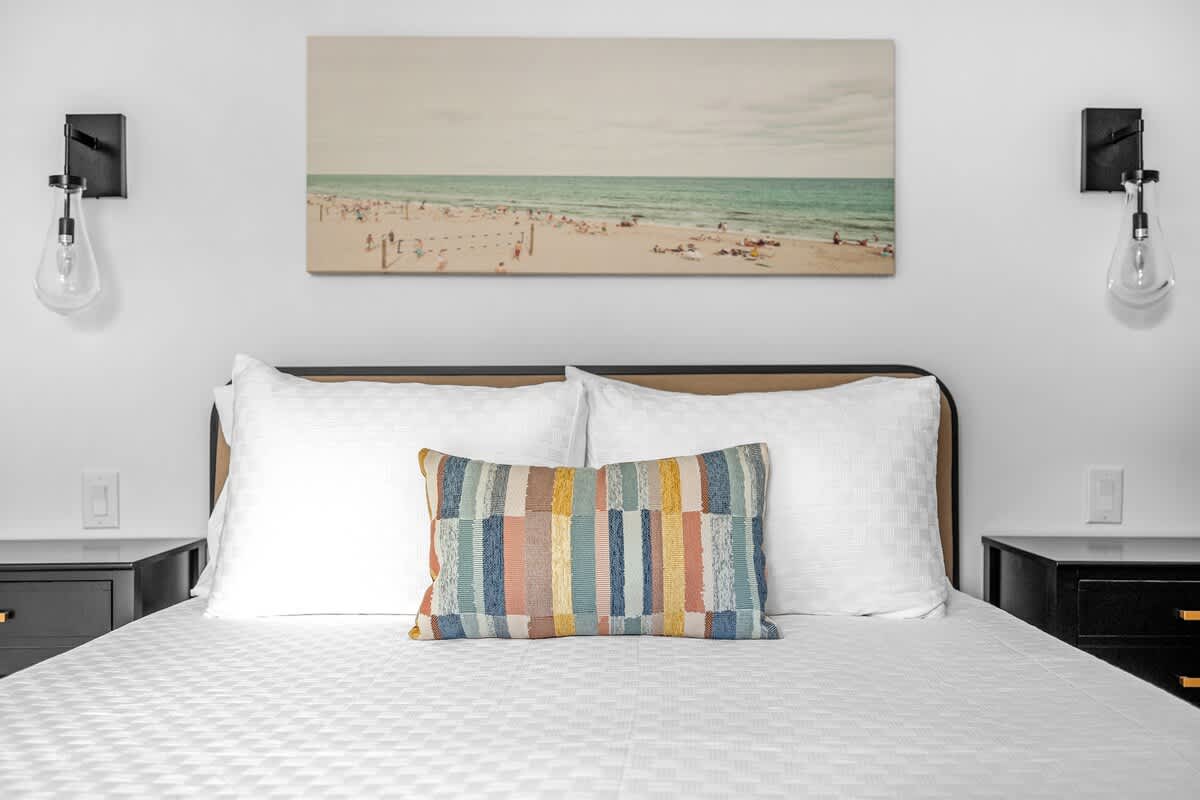 A close-up of the bed in a modern bedroom, featuring white bedding, a colorful striped accent pillow, and a large beach-themed artwork above the headboard. Black wall sconces are mounted on either side of the bed.
