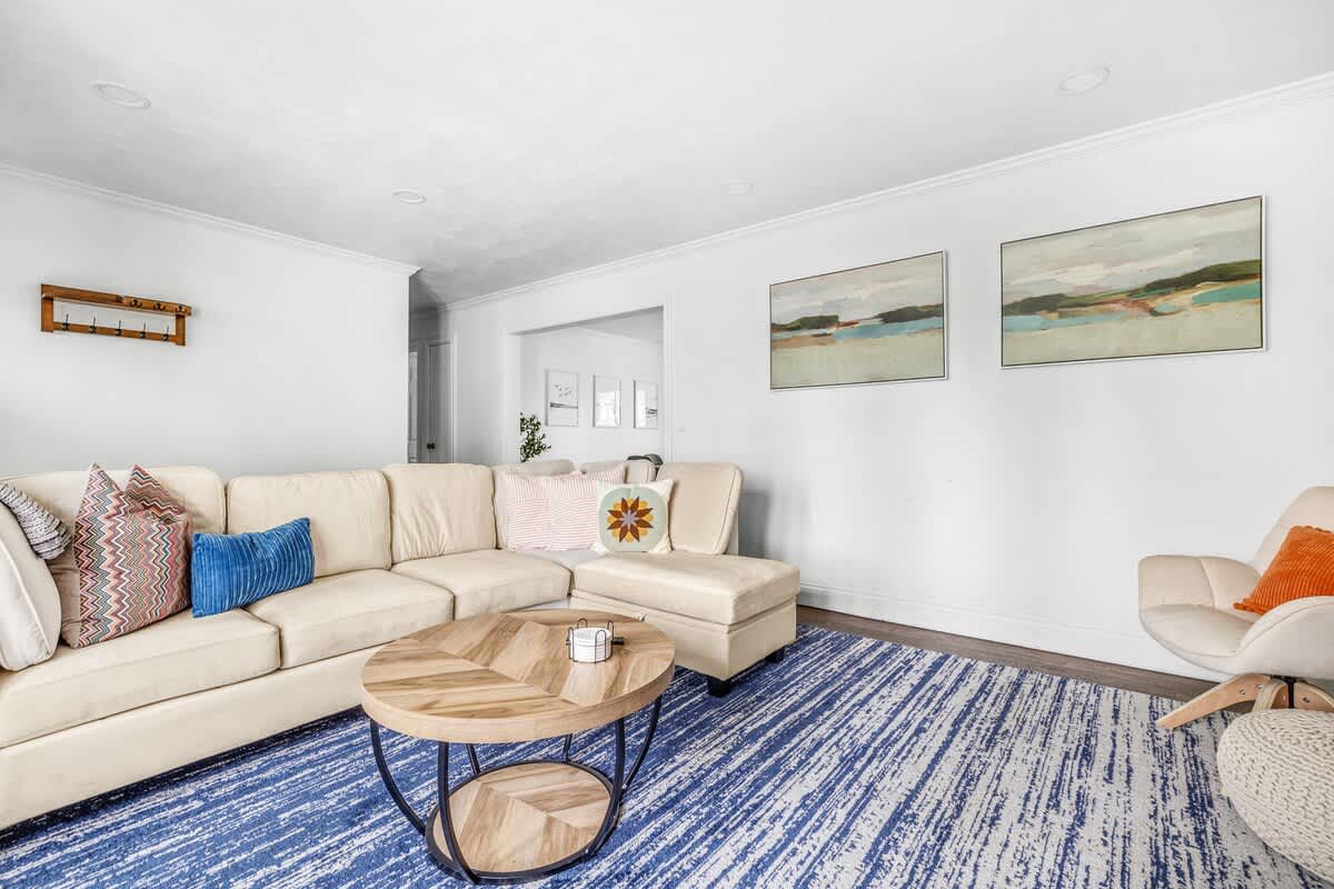 A second living area features a large cream sectional sofa with colorful throw pillows, a round wooden coffee table, and a blue and white striped rug, with two landscape paintings on the wall.