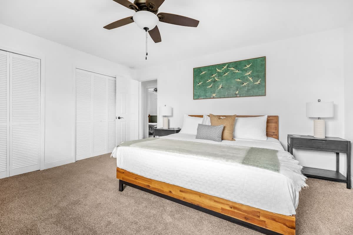 A bright bedroom with a wooden bed frame, white bedding, and a green and white throw blanket, flanked by dark nightstands with lamps. A large green artwork hangs above the bed, and white louvered closet doors are visible.