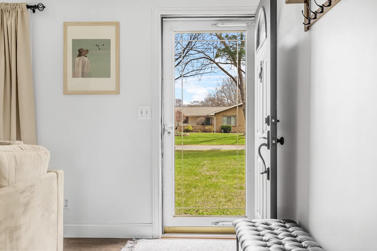 A bright entryway with a white door and a glass storm door opening to a view of a grassy front yard and a neighboring house. A framed artwork hangs on the white wall above a dark upholstered bench.
