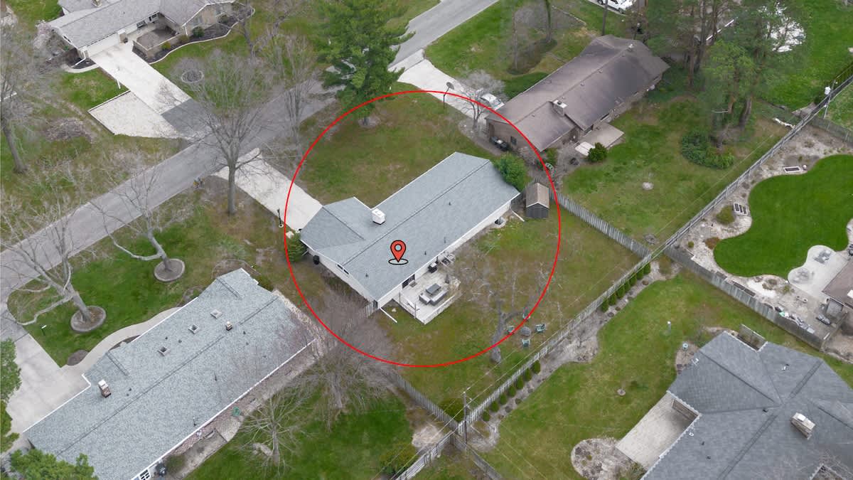 An aerial view of a single-story house with a gray roof, situated on a large grassy lot with mature trees. A red circle highlights the property, showing a deck in the backyard and surrounding residential properties.