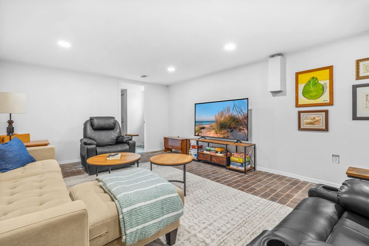 A cozy living room with a large flat-screen TV on a wooden media console, surrounded by board games. A beige sofa and black recliner provide seating on a patterned area rug, with brick-patterned flooring extending beyond the rug.