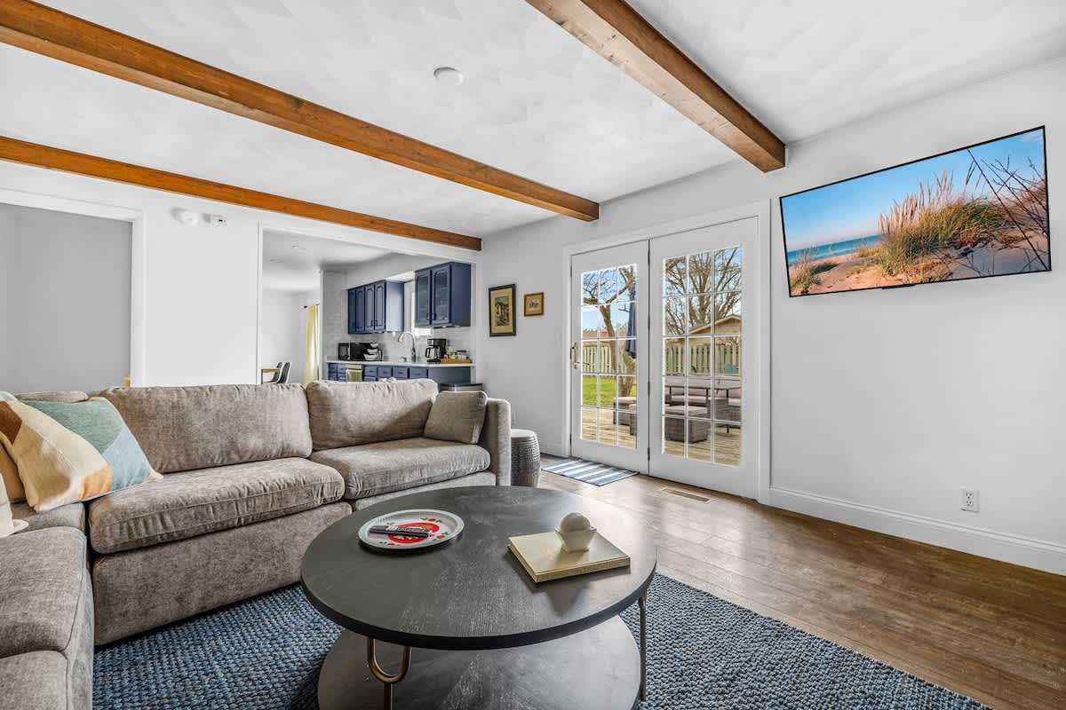 A cozy living room features a large grey sectional sofa, a round dark coffee table, and a blue patterned rug, with exposed wooden beams on the ceiling and French doors leading to a deck. A large TV is mounted on the white wall.