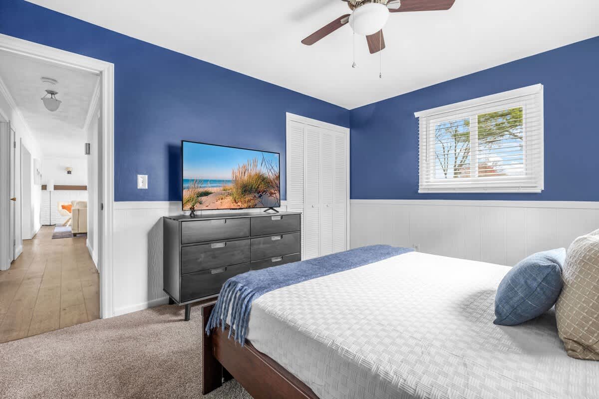 This bedroom features a dark blue accent wall with white wainscoting, a bed with white bedding, and a dark gray dresser with a large flat-screen TV. A ceiling fan and a window with white blinds are also visible, along with a white bi-fold closet door.