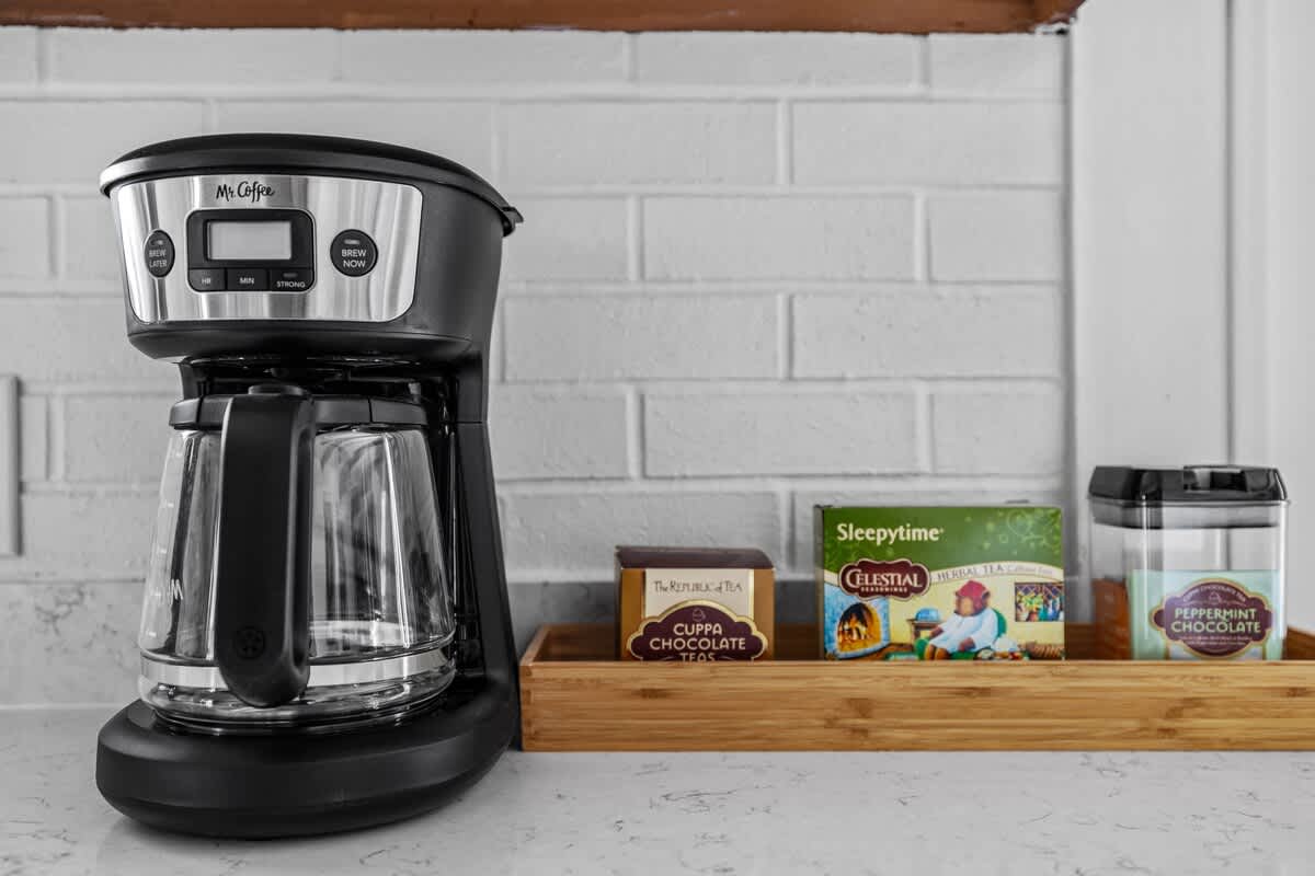A close-up of a coffee station on a white countertop, featuring a black Mr. Coffee maker, a wooden tray with various tea boxes, and a white subway tile backsplash.