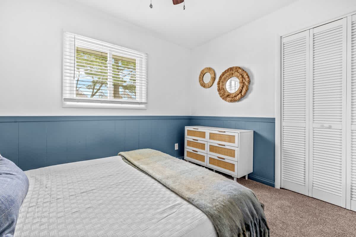 A bedroom with a gray bed and white bedding is shown, featuring a white wall with blue wainscoting and a white dresser with woven drawers. A window with white blinds and a white bi-fold closet door are also visible.