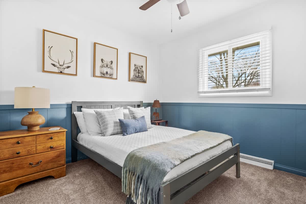 This bedroom features a gray bed with white bedding and a blue throw blanket, set against a white wall with blue wainscoting. A wooden dresser with a lamp is on the left, and a window with white blinds allows natural light.
