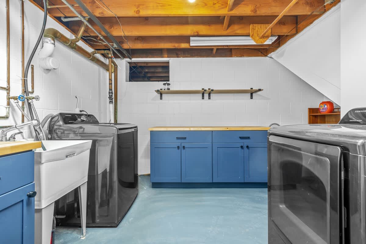 A functional laundry room with exposed wooden beams and pipes on the ceiling, white cinder block walls, and a blue painted concrete floor. It features a washing machine, dryer, utility sink, and blue base cabinets with wooden countertops.