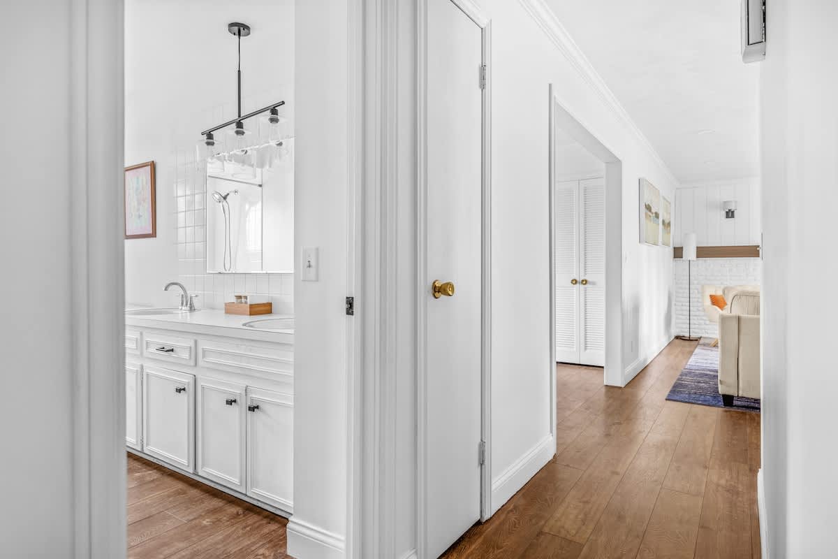 A hallway with hardwood floors and white walls leads past a closed door with a brass knob, offering a glimpse into a bathroom on the left and a living area with a fireplace and blue rug further down the hall.