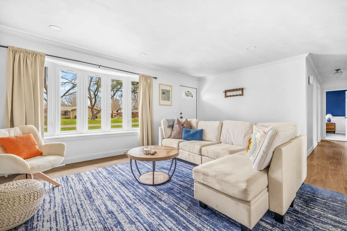 A spacious living room features a cream sectional sofa with colorful throw pillows, a large bay window, and a round wooden coffee table on a blue and white striped area rug.
