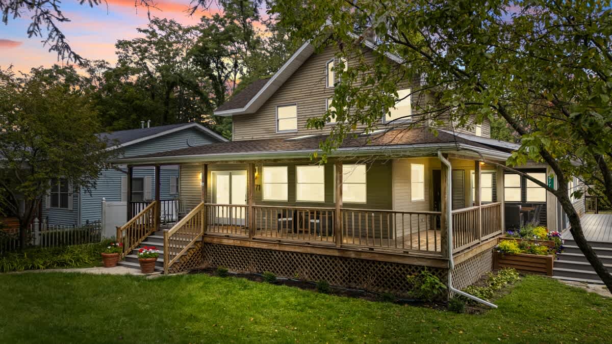 The front exterior of a two-story house with light brown siding and a wrap-around wooden porch, illuminated by interior lights at dusk, surrounded by green grass and trees.