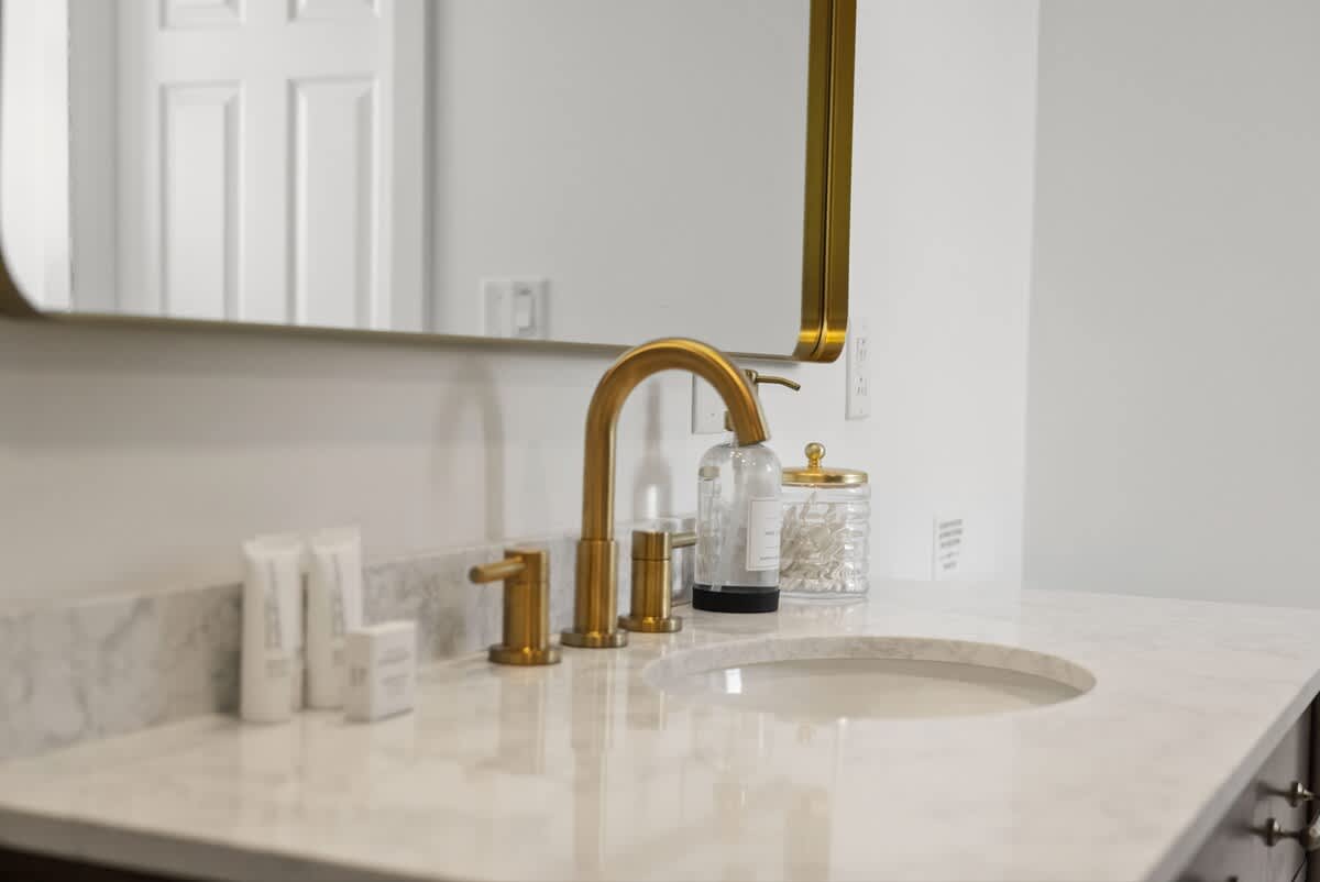Close-up of a bathroom vanity featuring a white marble countertop, a modern gold faucet, and a rectangular sink, with various toiletries neatly arranged.