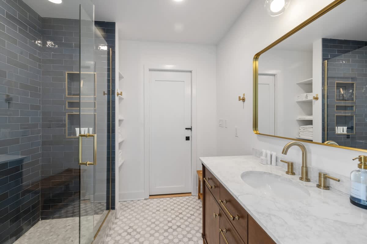 Wide shot of the modern bathroom, showcasing the dark wood vanity with a white marble top, gold fixtures, a large rectangular mirror, and the walk-in shower with dark grey subway tiles and glass enclosure.