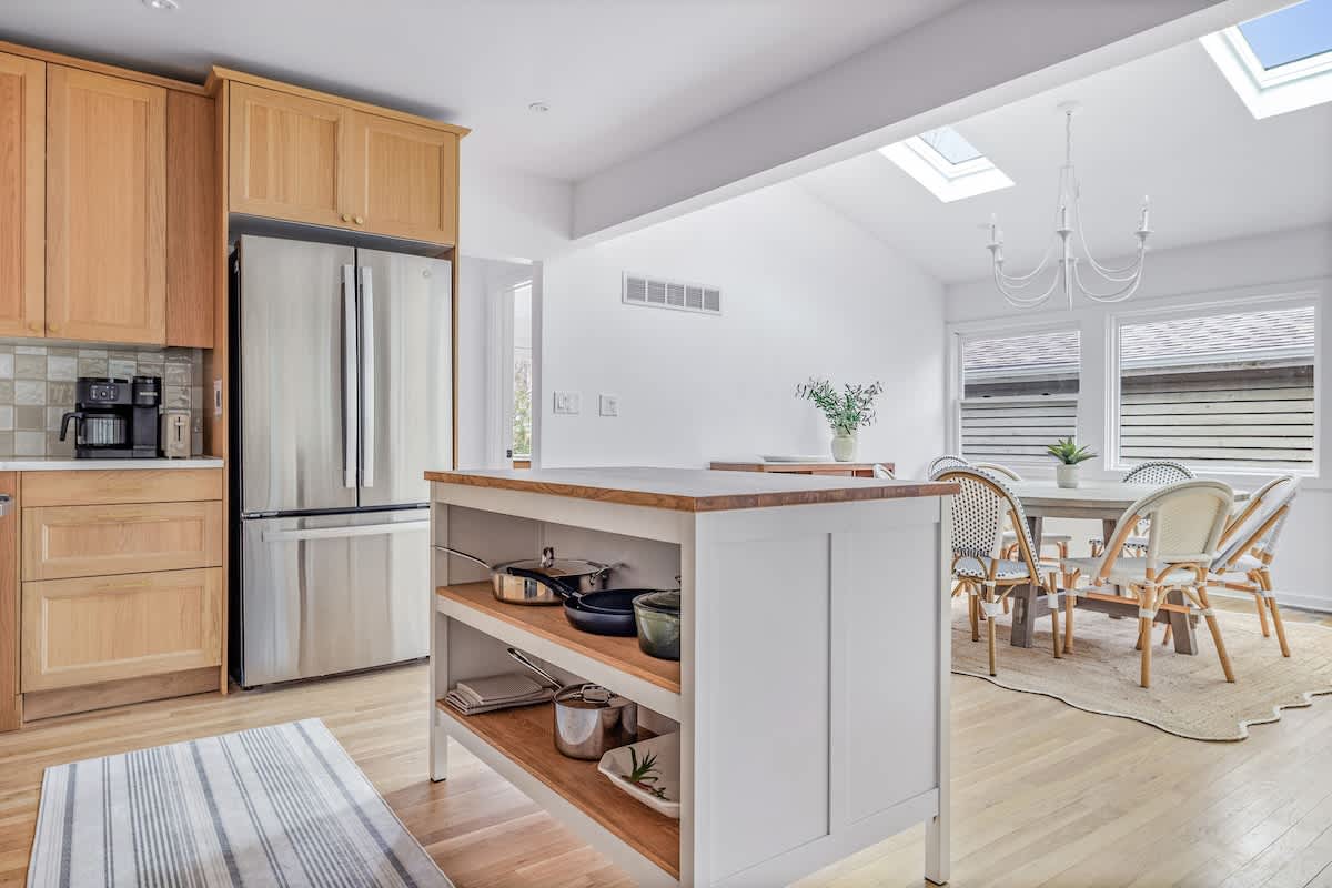 View of the kitchen and adjacent dining area, featuring light wood cabinets, a stainless steel refrigerator, and a white kitchen island with open shelving. The dining area has a round table and wicker chairs under a skylight.