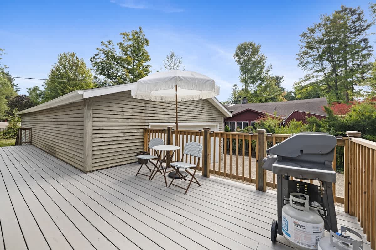 A spacious light grey composite deck features a BBQ grill, a white patio umbrella, and a small table with two chairs, adjacent to a detached garage with light brown siding.