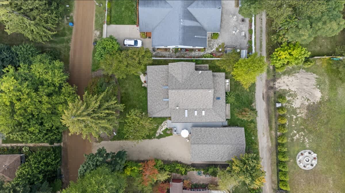 An aerial view of the property showcasing the main house with a multi-gabled roof, a detached garage, and a gravel driveway, surrounded by lush green trees and neighboring properties.