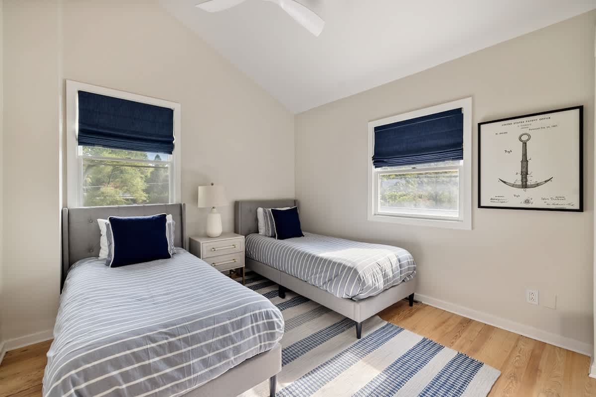 Bedroom with two twin beds featuring grey upholstered frames and blue and white striped bedding. Light hardwood floors, a blue and white striped rug, and two windows with navy blue Roman shades create a cohesive nautical theme.
