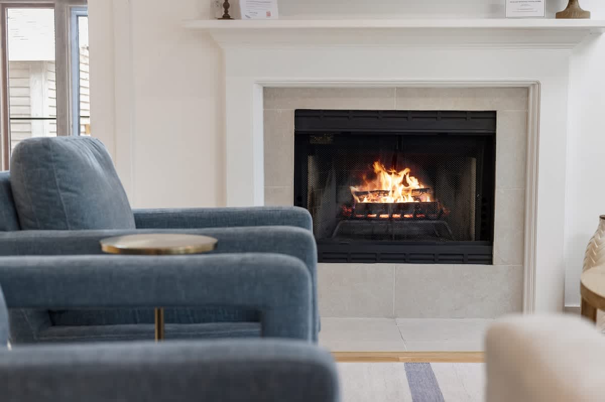 Cozy living room featuring a white stone fireplace with a black insert and burning logs, flanked by light blue upholstered armchairs with brass accents. The room has light hardwood floors and a simple white mantel.