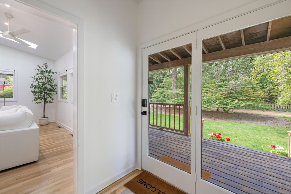 Bright entryway with a glass door leading to a wooden porch and a view of the green yard. Hardwood floors extend into an adjacent living area with a white sofa and a potted tree.