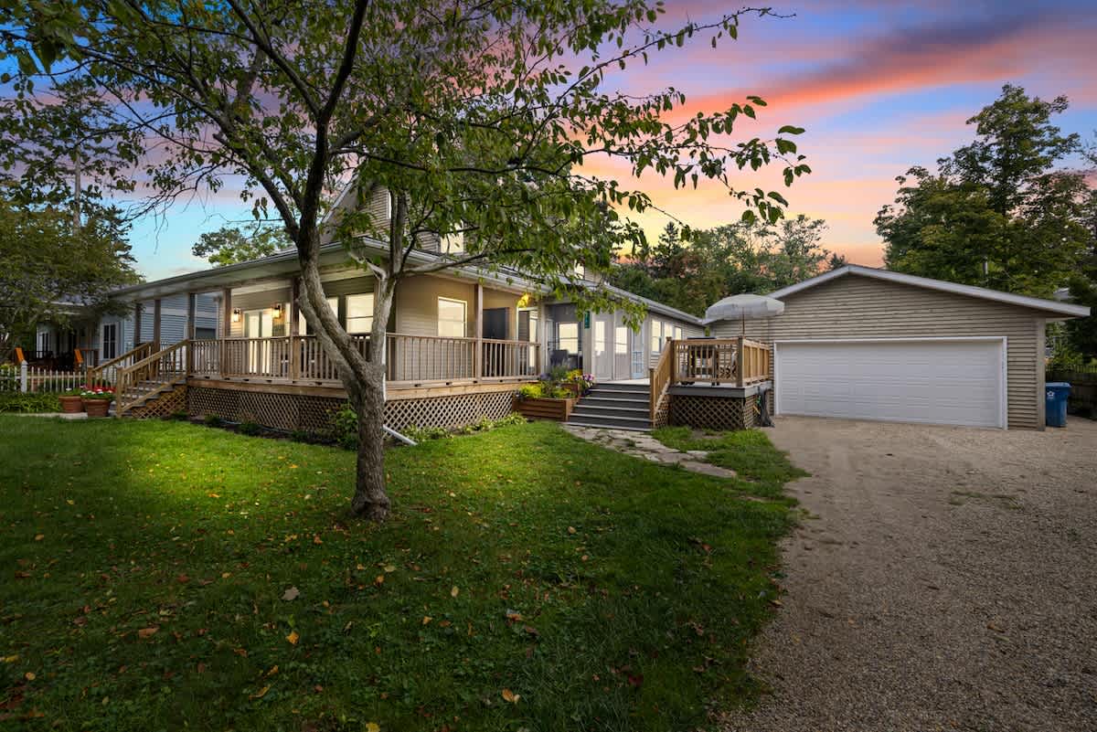 A wide exterior view of the two-story house with a wrap-around porch and a detached garage with a white door, set against a vibrant sunset sky and surrounded by trees and a gravel driveway.