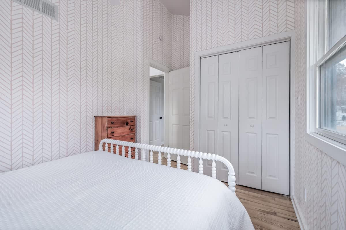 View of the bedroom from a different angle, showcasing the light hardwood floors and herringbone wallpaper. A white spindle bed is visible, along with a wooden dresser and a white bi-fold closet door.