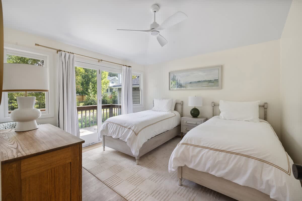 Bright bedroom featuring two twin beds with white bedding, a wooden dresser, and a ceiling fan, with large glass doors opening to an outdoor deck.