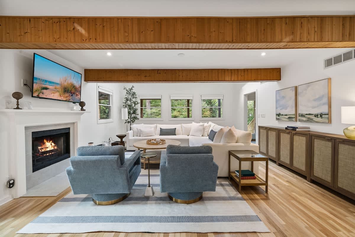 Spacious living room with exposed wooden beams, a white sectional sofa, and two blue swivel armchairs. A large TV is mounted above a white fireplace, and a long wooden console with woven doors lines the far wall.