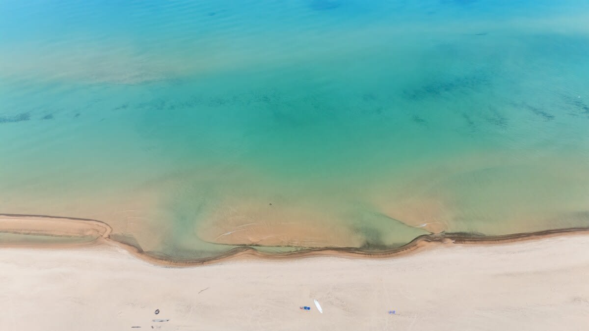 An aerial view of a serene sandy beach meeting clear turquoise ocean water, with gentle waves creating subtle patterns on the shallow seabed.