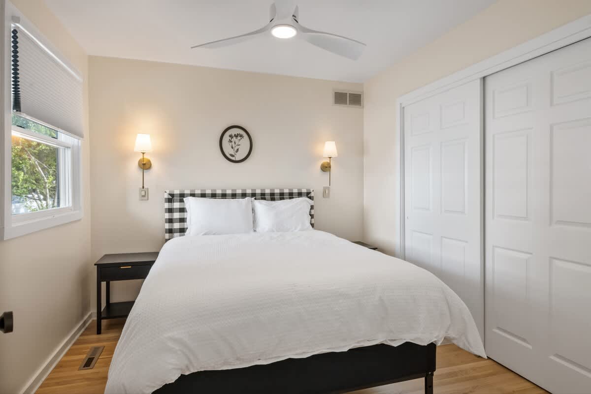 Full view of the bedroom with a queen bed featuring a plaid headboard, a black nightstand, a window with blinds, and a white bi-fold closet door. A ceiling fan is also visible.
