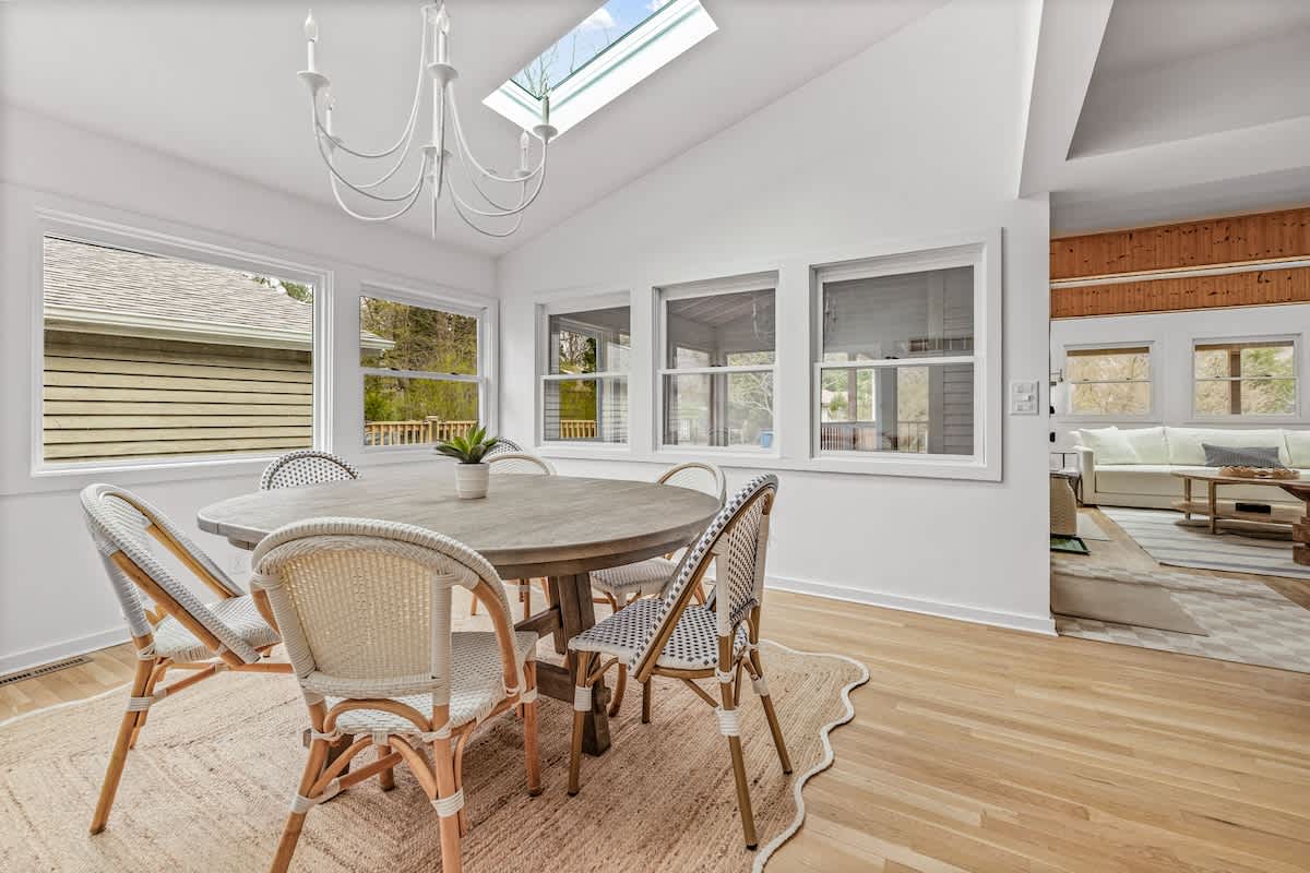 Bright dining room with a light wood oval dining table surrounded by white and black wicker chairs, set on a textured rug. Large windows offer views of the outdoors, and a white chandelier hangs from the vaulted ceiling with a skylight.