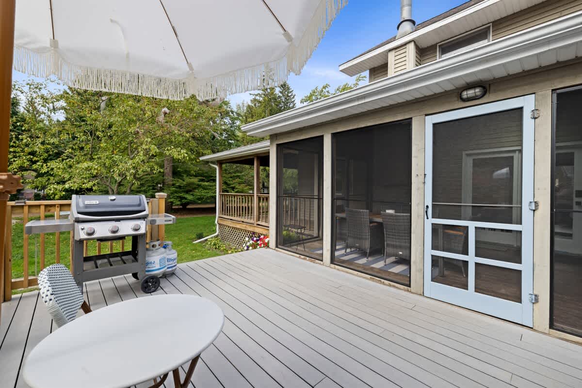A light grey composite deck with a BBQ grill and a white patio umbrella, offering access to a screened porch with a light blue door, surrounded by lush green trees.