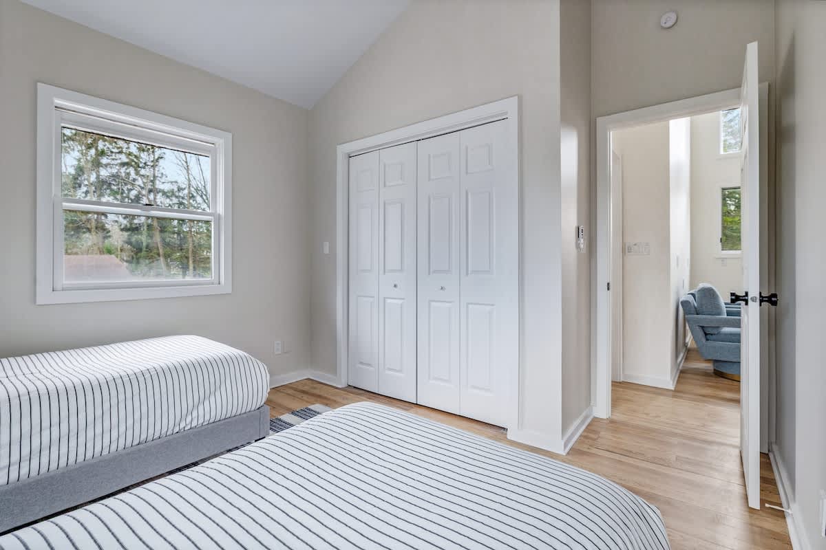 Another view of the bedroom with two twin beds, showing a white bi-fold closet door and a large window. The light hardwood floors and neutral wall color are consistent with the room's design.