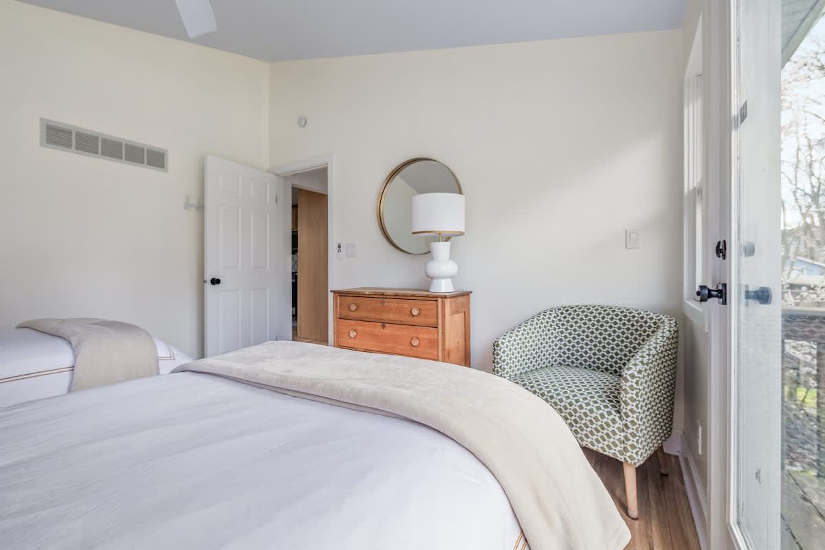 Another view of the bright bedroom, highlighting one of the twin beds with white bedding, a patterned accent chair, a wooden dresser with a round mirror, and a glass door leading outside.