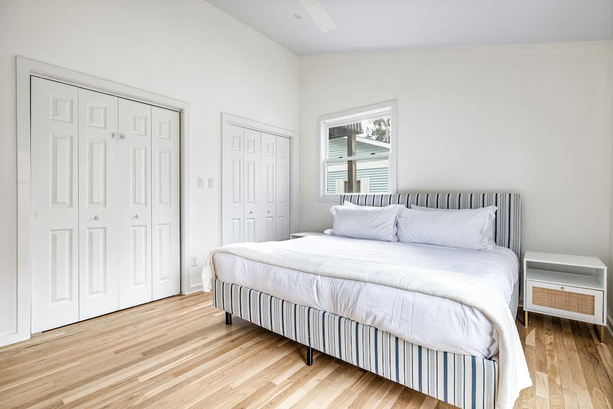 Bright bedroom with light hardwood floors and white walls, featuring a queen-sized bed with a blue and white striped upholstered frame and white bedding. Two white closets with paneled doors are visible, along with a modern nightstand.