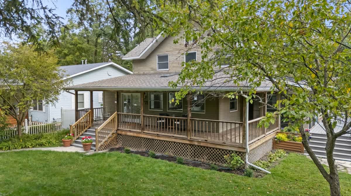 The front exterior of a two-story house with light brown siding and a wrap-around wooden porch with stairs, surrounded by a green lawn and mature trees under a bright sky.