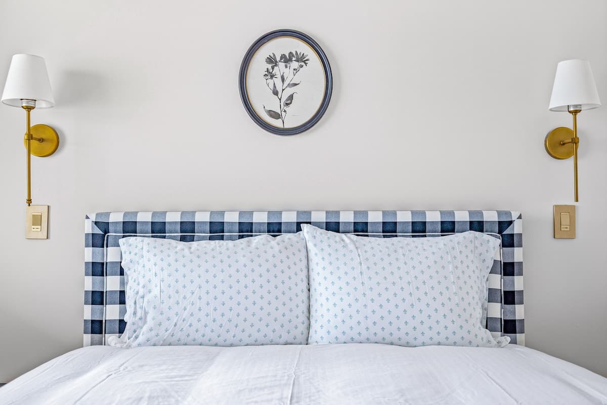 Cozy bedroom featuring a bed with a distinctive blue and white plaid upholstered headboard, flanked by two gold wall sconces, and a circular botanical art piece above the bed.
