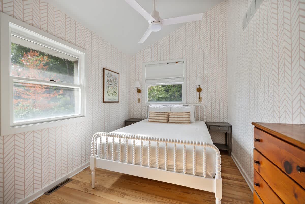 Cozy bedroom with light hardwood floors and walls covered in a subtle herringbone pattern wallpaper. A white spindle bed with white bedding and striped accent pillows is centered between two nightstands with wall-mounted sconces.