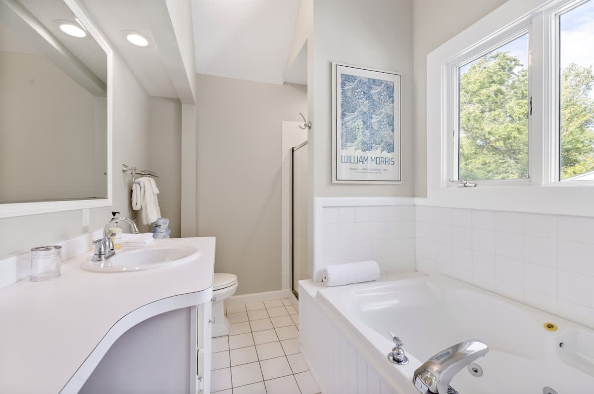 Bathroom with a white jetted tub, white subway tile wainscoting, and a vanity with a white countertop and sink. A framed William Morris print hangs on the wall, and a glass-enclosed shower is visible in the background.