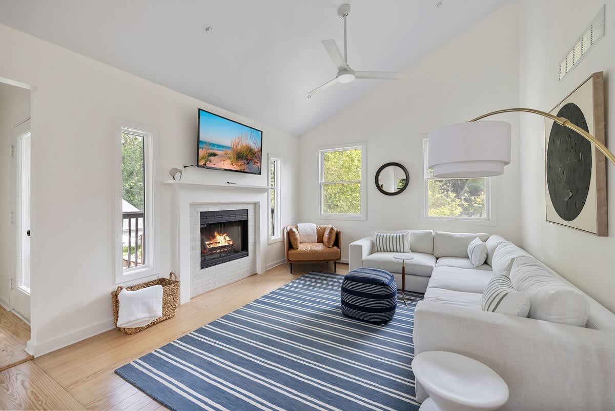 Spacious living room featuring a large white sectional sofa with striped pillows, a blue and white striped area rug, and light hardwood floors. A wall-mounted TV is above a white fireplace, and a modern ceiling fan hangs from the vaulted ceiling.